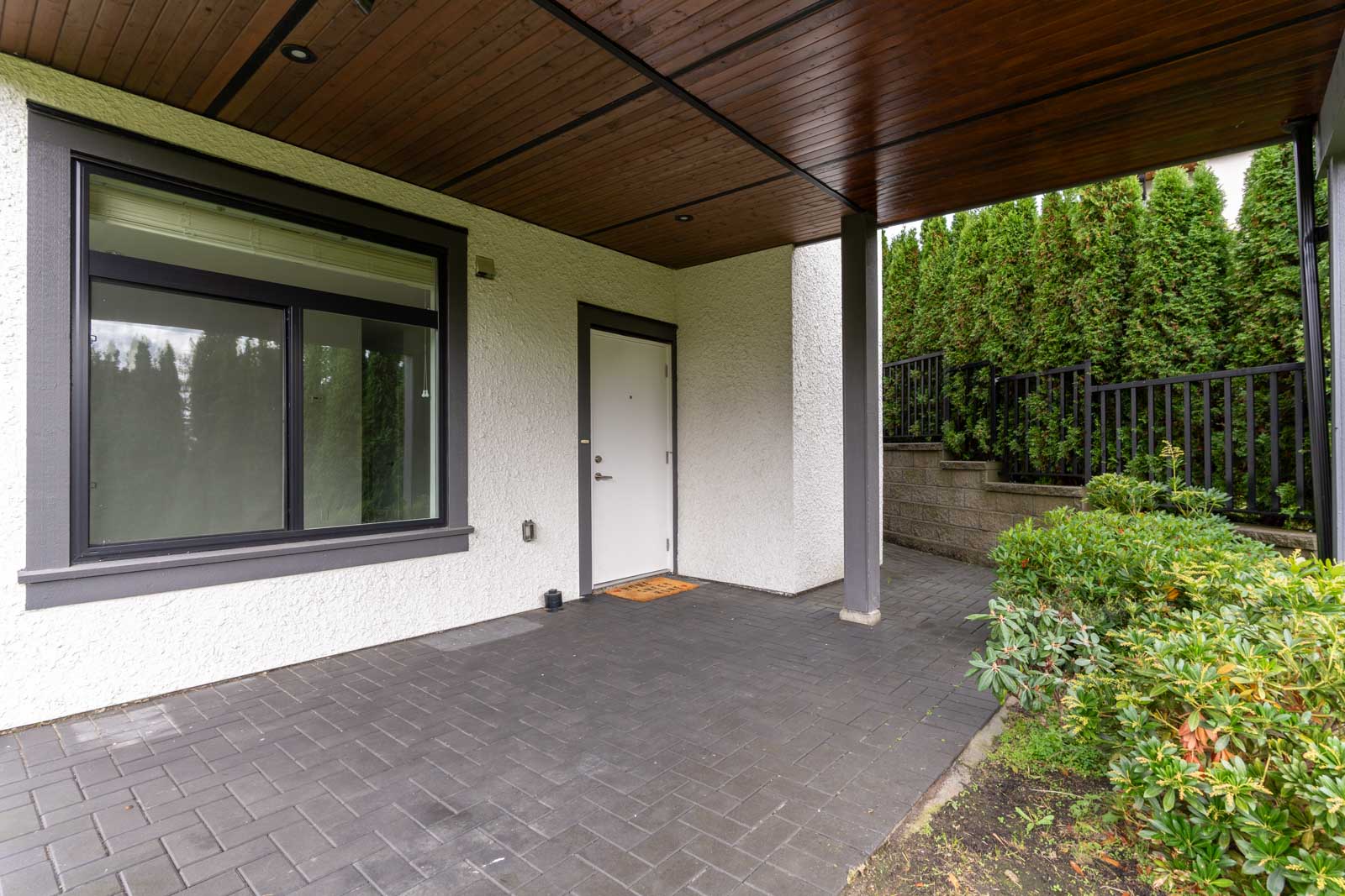 Covered patio area with black tile flooring, white door, large window, and surrounding greenery next to a black metal fence.