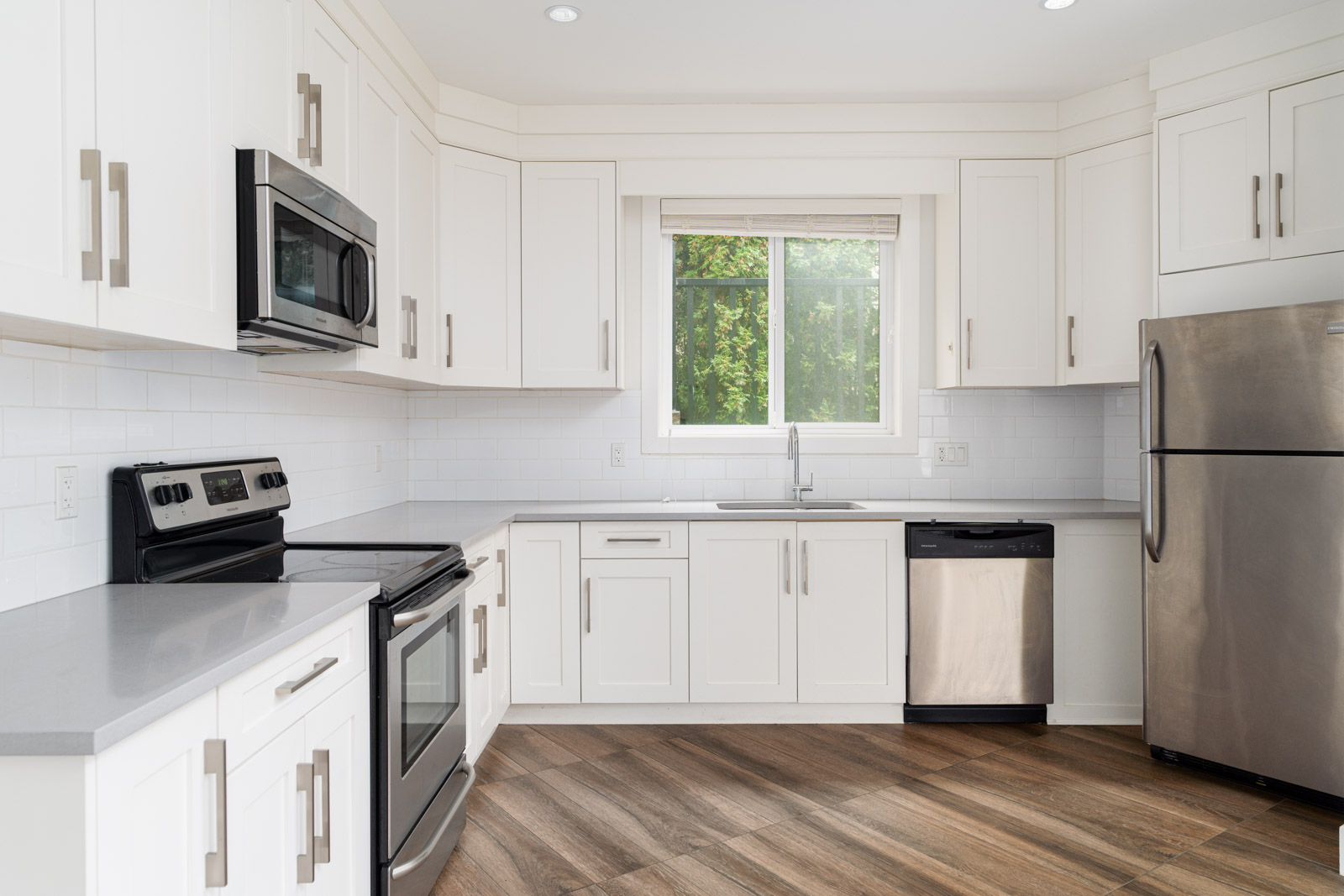 Modern kitchen with white cabinets, stainless steel appliances, a large window above the sink, and wood-patterned flooring.