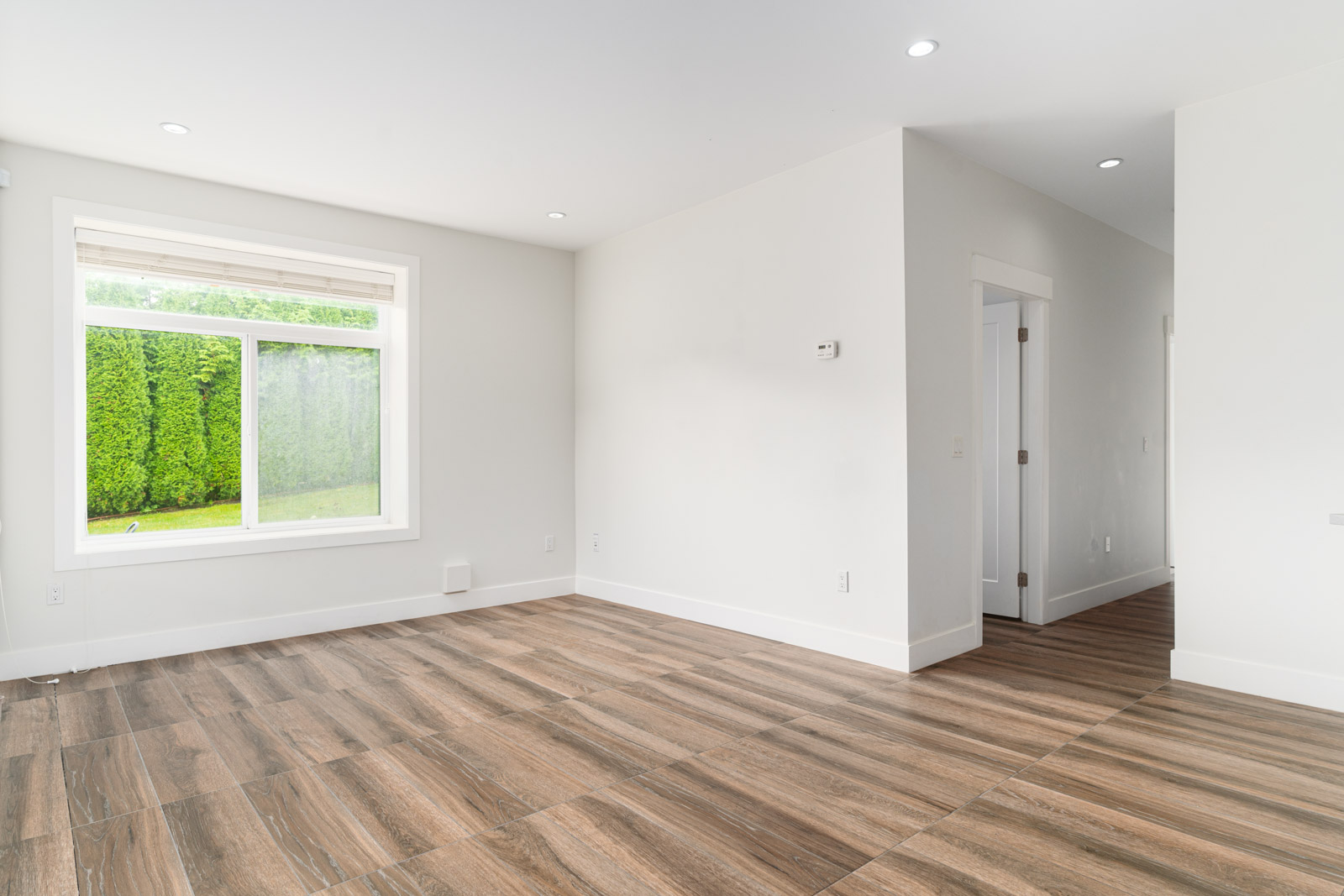 Empty room with white walls, large window showing greenery outside, and wood-patterned tile flooring; hallway visible on the right.