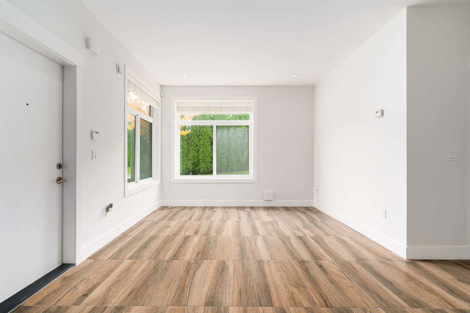 Empty, unfurnished room with white walls, large windows, wood-look tile flooring, and a closed white door on the left. Natural light enters through the windows.