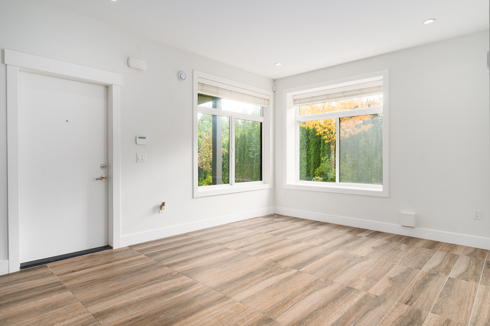 Empty room with large windows, white walls, wood-look tile flooring, and a closed white door; green trees and autumn foliage visible outside.