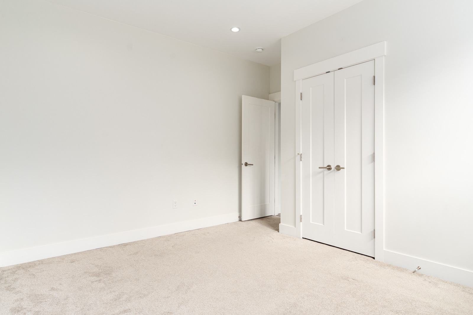 Empty room with beige carpet, white walls, a closed double-door closet, and an open door leading to another space. Ceiling lights are on.