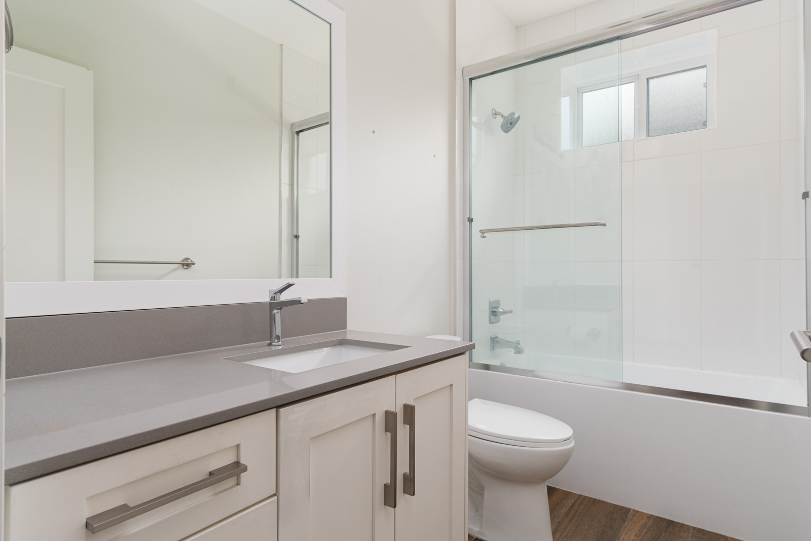 Modern bathroom with a gray countertop, white vanity, large mirror, toilet, and bathtub with a glass sliding door and window above.