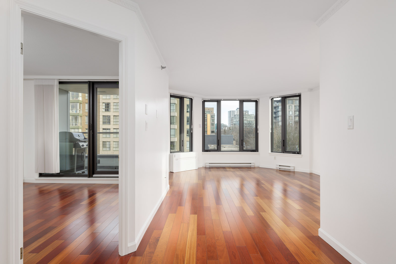 Bright, empty apartment with polished wooden floors, large windows, and a balcony with city views. White walls and natural light fill the open living area.