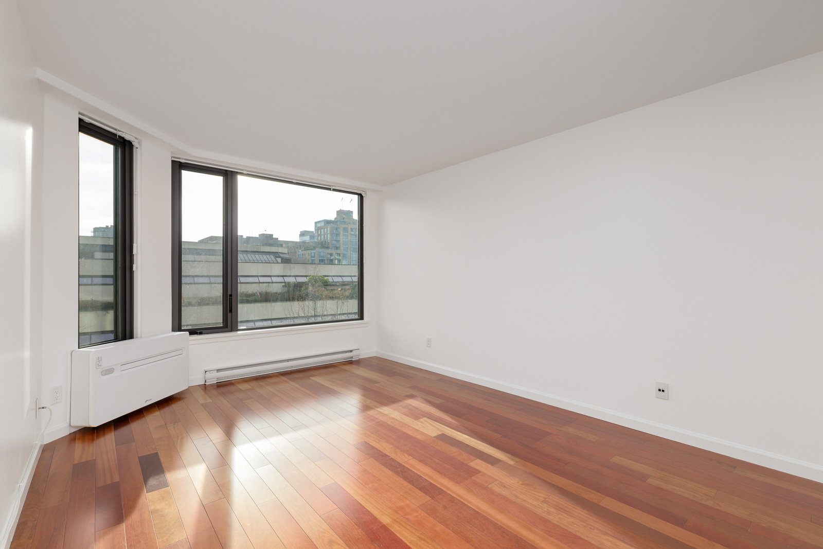 Empty room with white walls, large windows, and hardwood floors. A wall-mounted air conditioning unit is visible below the windows, with a view of buildings outside.