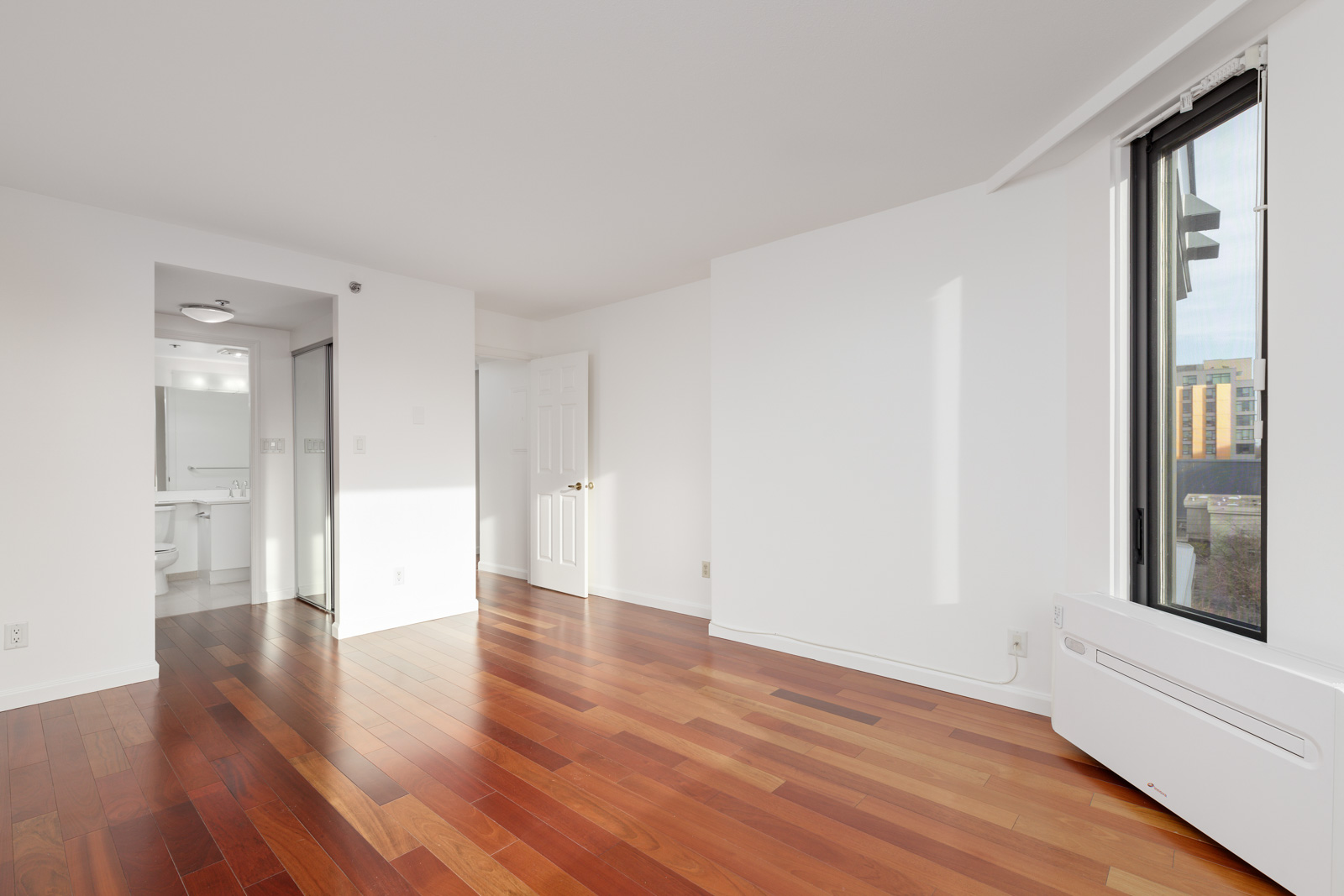 Bright, empty room with hardwood floors, white walls, large window, and an open doorway leading to a bathroom with a mirror and toilet visible.