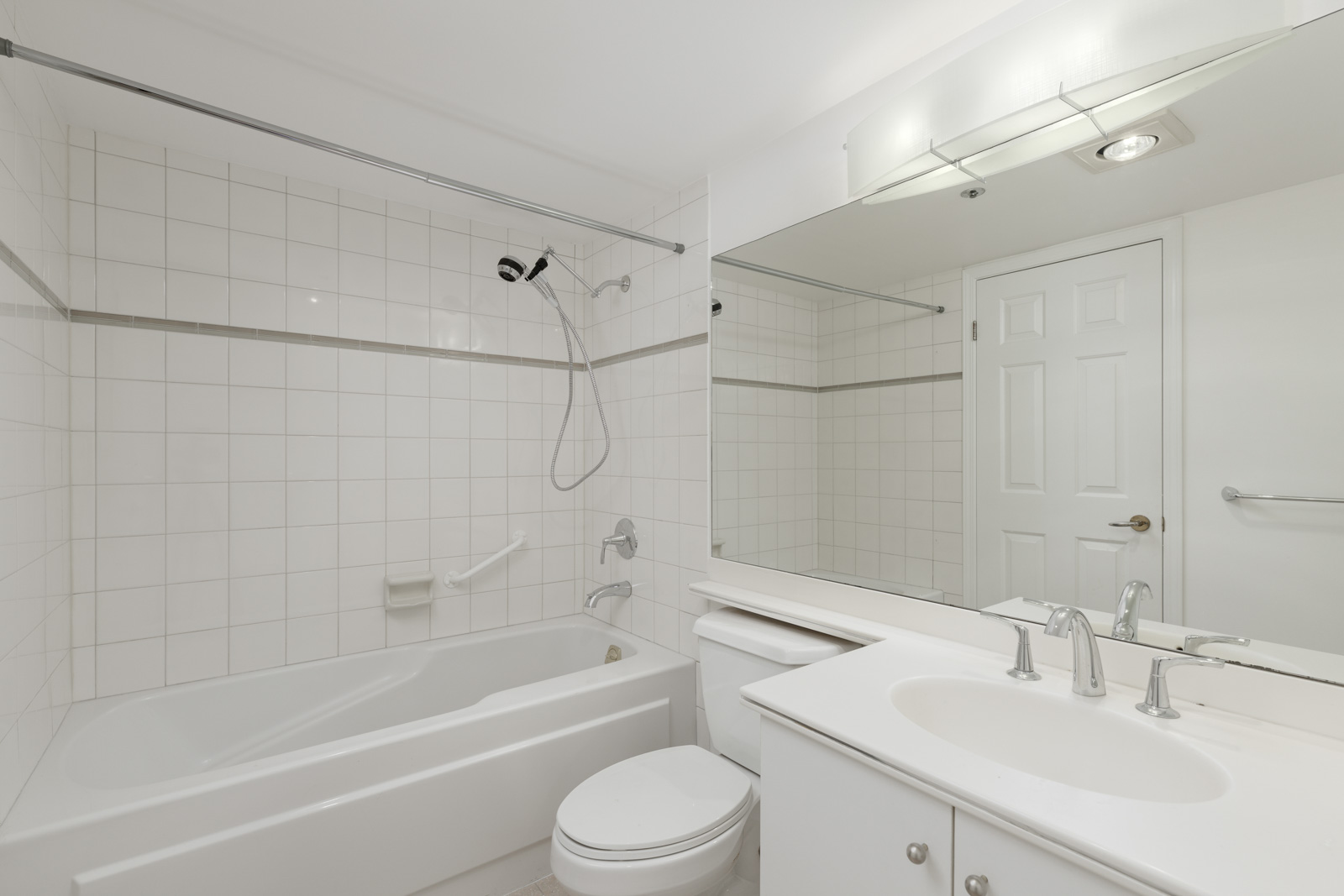 White bathroom with a bathtub and shower, toilet, large mirror, sink with cabinet, and overhead lighting. White tile covers the shower area and walls.