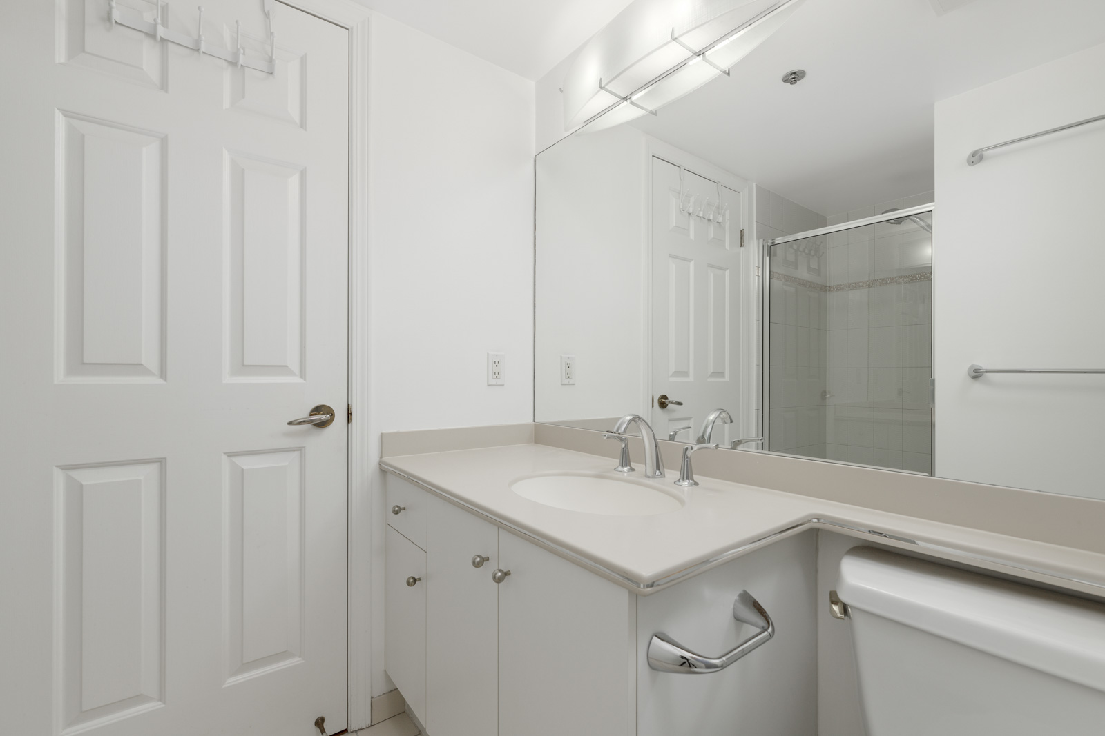 White bathroom with a double sink vanity, large mirror, overhead light, towel rack, and a shower with glass doors.