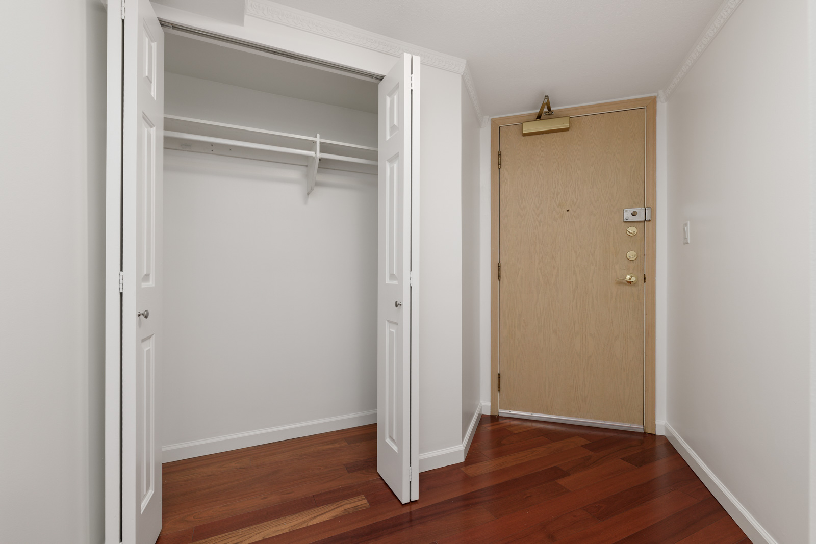 A small entryway with a light wood front door and an open closet with double white doors, a shelf, and a hanging rod. The floor is dark wood.
