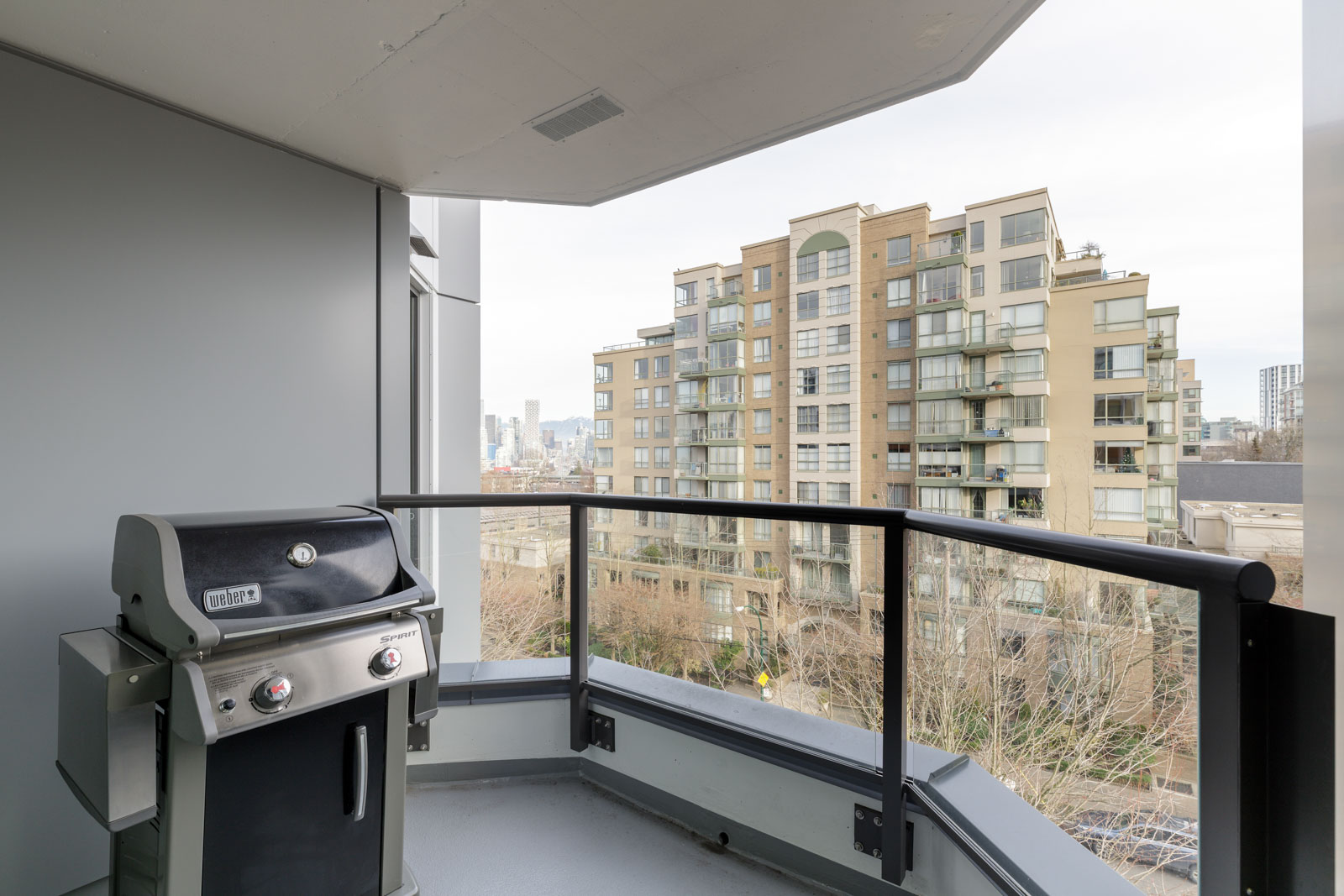 A gas grill on a high-rise balcony with a glass railing, overlooking nearby apartment buildings and a distant city skyline.