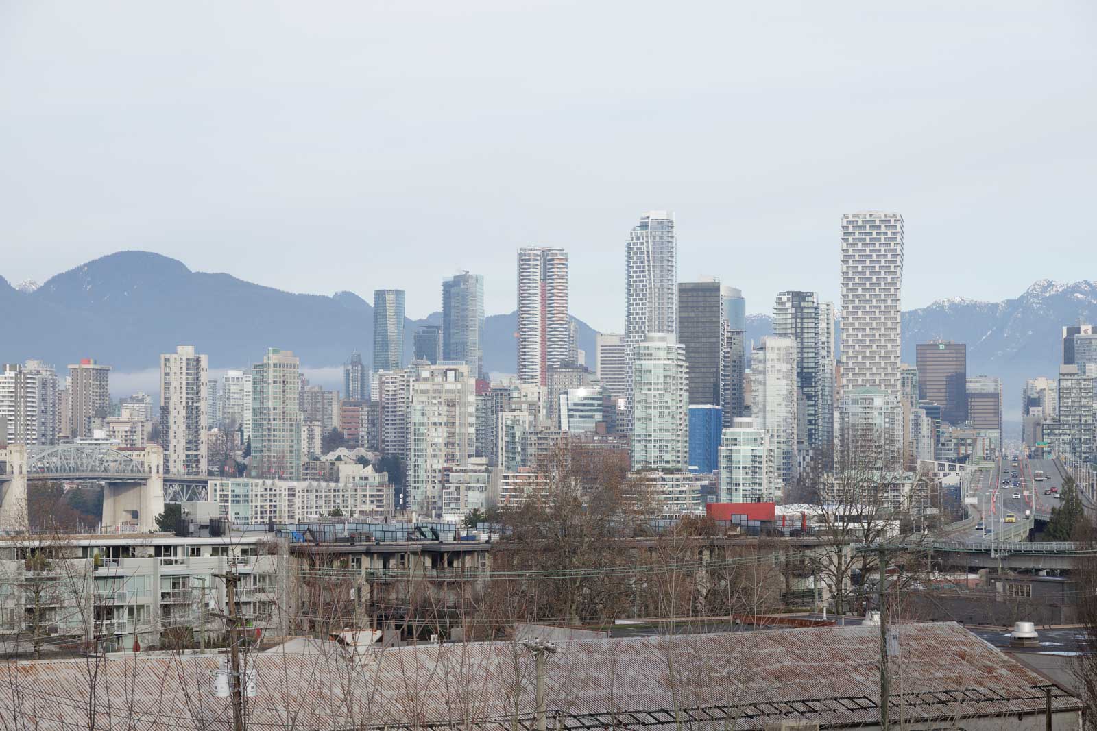 A city skyline with numerous high-rise buildings set against a backdrop of mountains under an overcast sky. In the foreground, there are lower buildings and power lines.