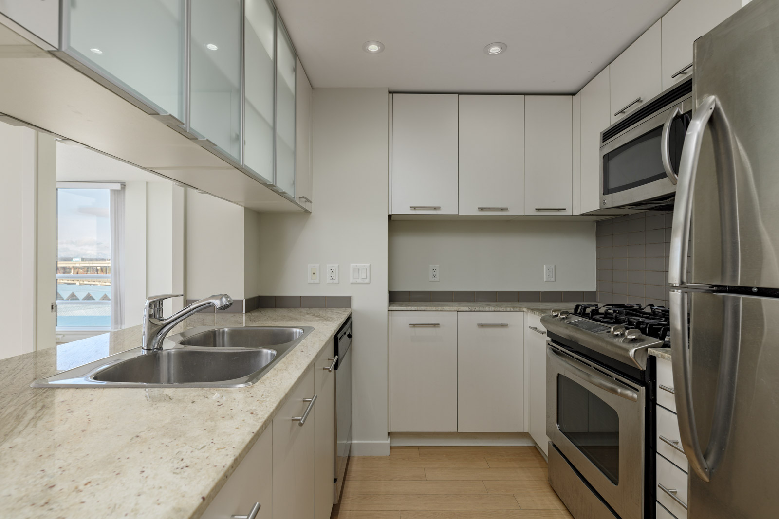 Modern kitchen with light-colored cabinets, stainless steel appliances, double sink, granite countertops, and a gas stove; window view in the background.