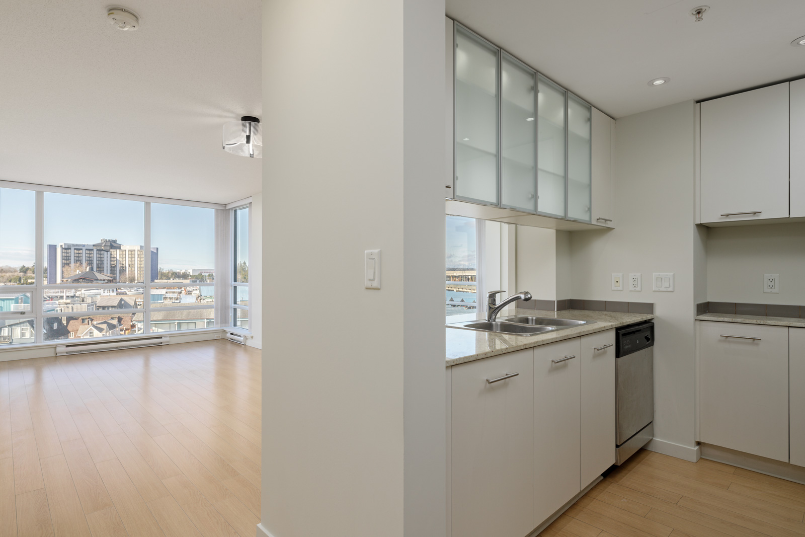 Modern apartment interior showing a kitchen with white cabinets and a dishwasher on the right, and a bright living area with large windows and city views on the left.