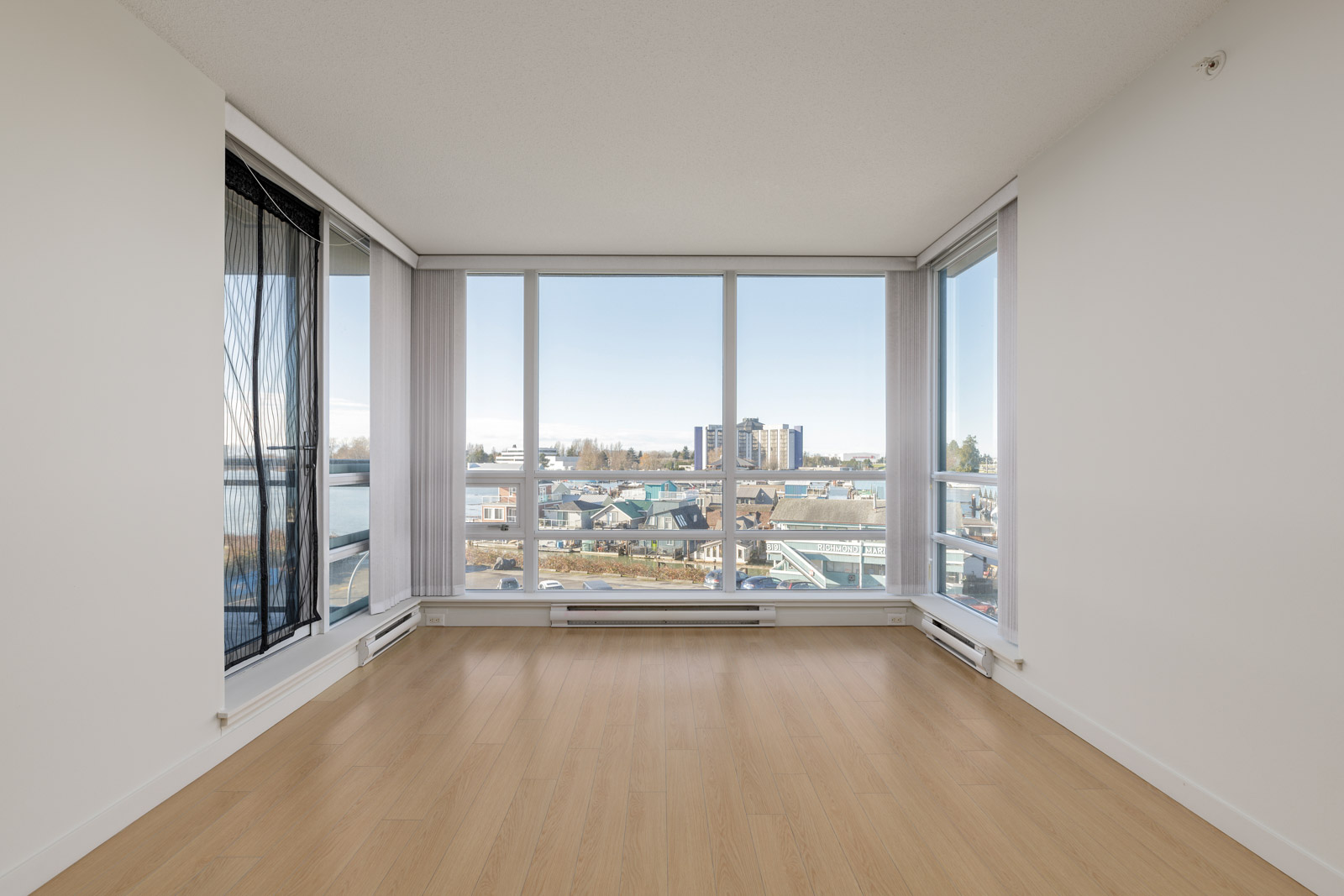 Empty bright room with light wood flooring, white walls, and large floor-to-ceiling windows overlooking a cityscape and distant water.