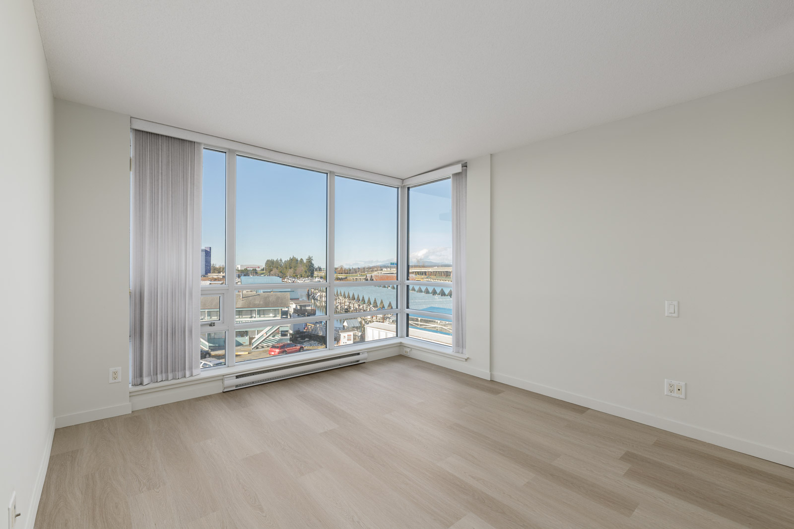 Unfurnished room with light wood flooring, large corner windows, white walls, and a view of water and buildings outside.