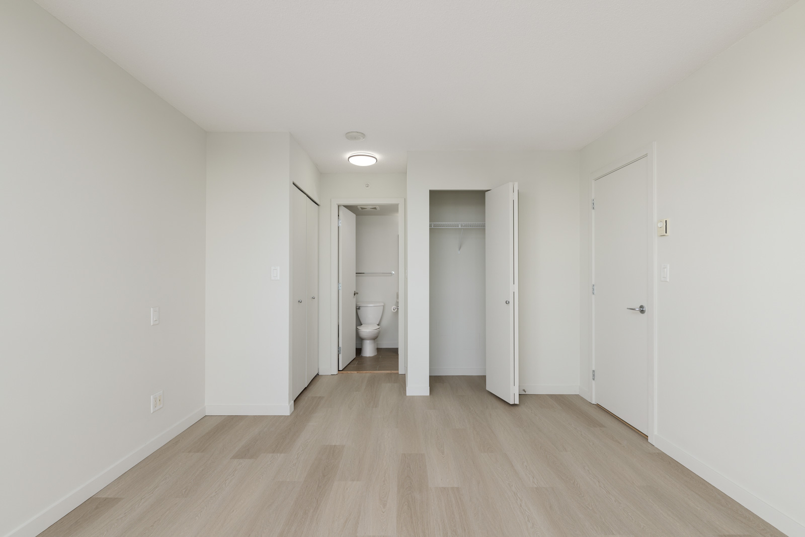 Empty room with light wood flooring, an open closet, a door, and an open doorway leading to a bathroom with visible toilet. Walls are white and the space is well-lit.