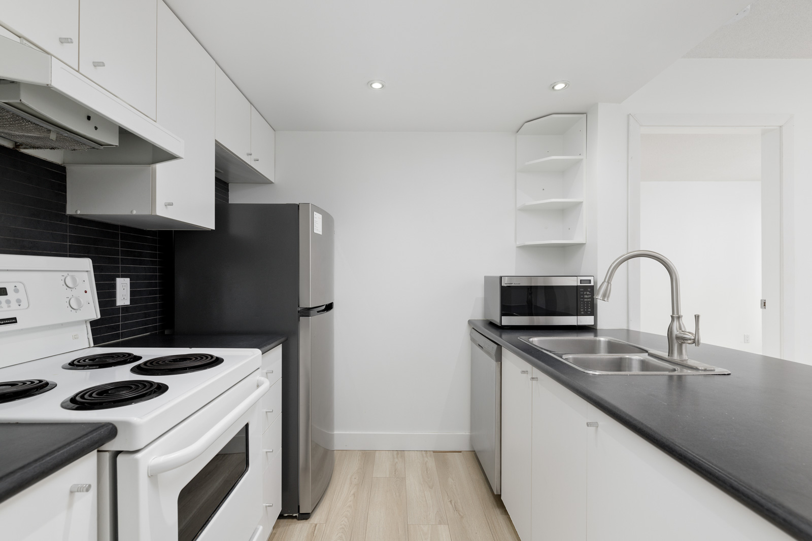 Modern kitchen with white cabinets, black countertops, electric stove, refrigerator, microwave, sink, and light wood flooring.