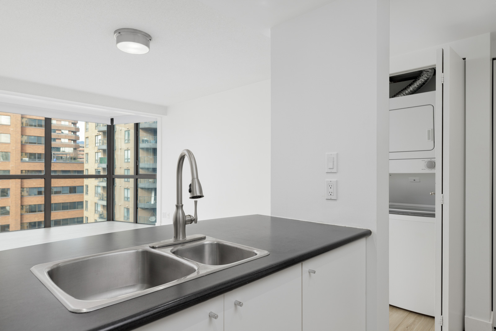 Modern kitchen with a double sink and black countertop, large windows with city view, and a stacked washer and dryer in a closet.