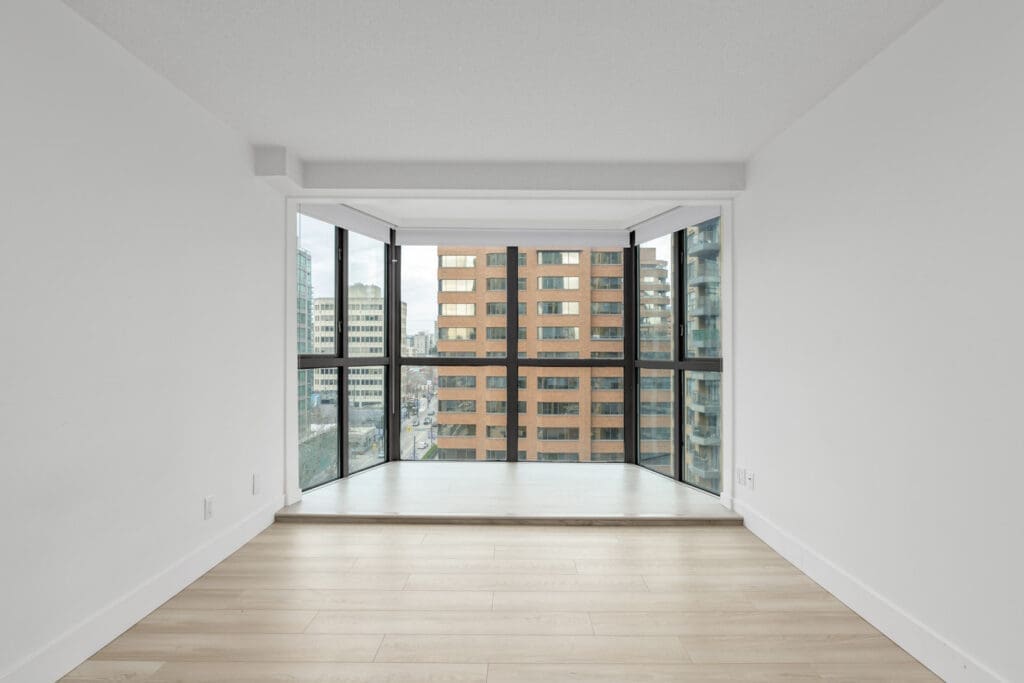 Empty room with light wood flooring, white walls, and a floor-to-ceiling corner window overlooking city buildings.