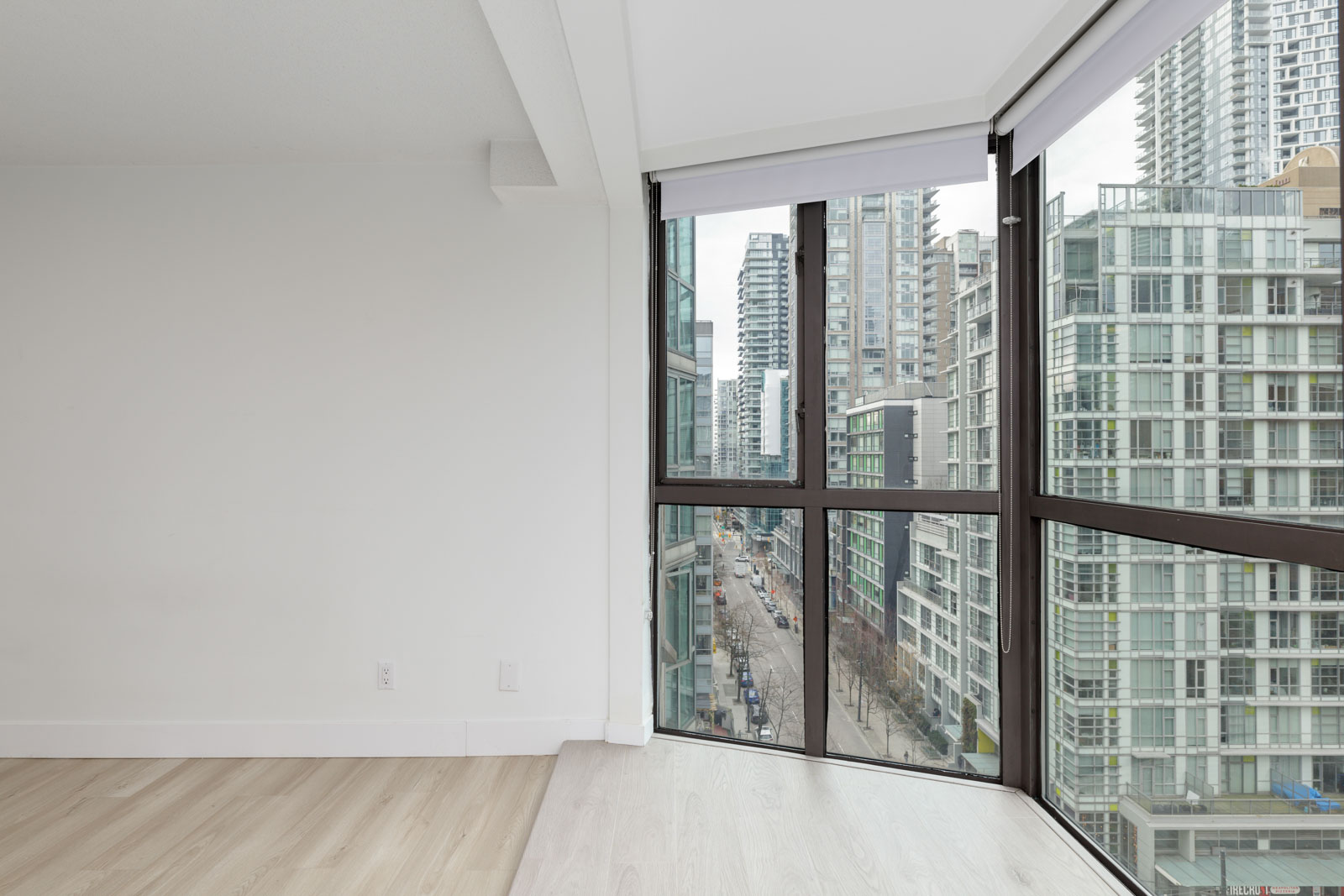 Corner of a modern, unfurnished apartment with large floor-to-ceiling windows overlooking a city street and high-rise buildings.