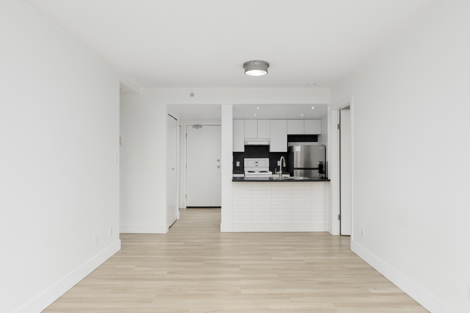 Unfurnished modern apartment interior with light wood flooring, white walls, and an open kitchen featuring stainless steel appliances and white cabinetry.