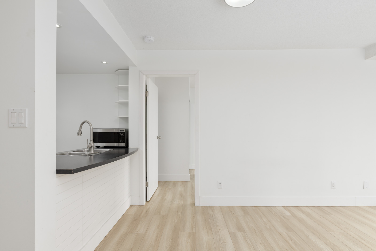 Minimalist kitchen with a sink, microwave, open shelves, and a black countertop connected to a bright, empty room with light wood flooring and white walls.