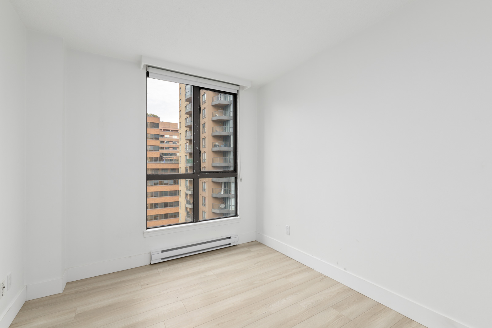 Empty white room with light wood flooring, a large window with a view of nearby apartment buildings, and a baseboard heater below the window.