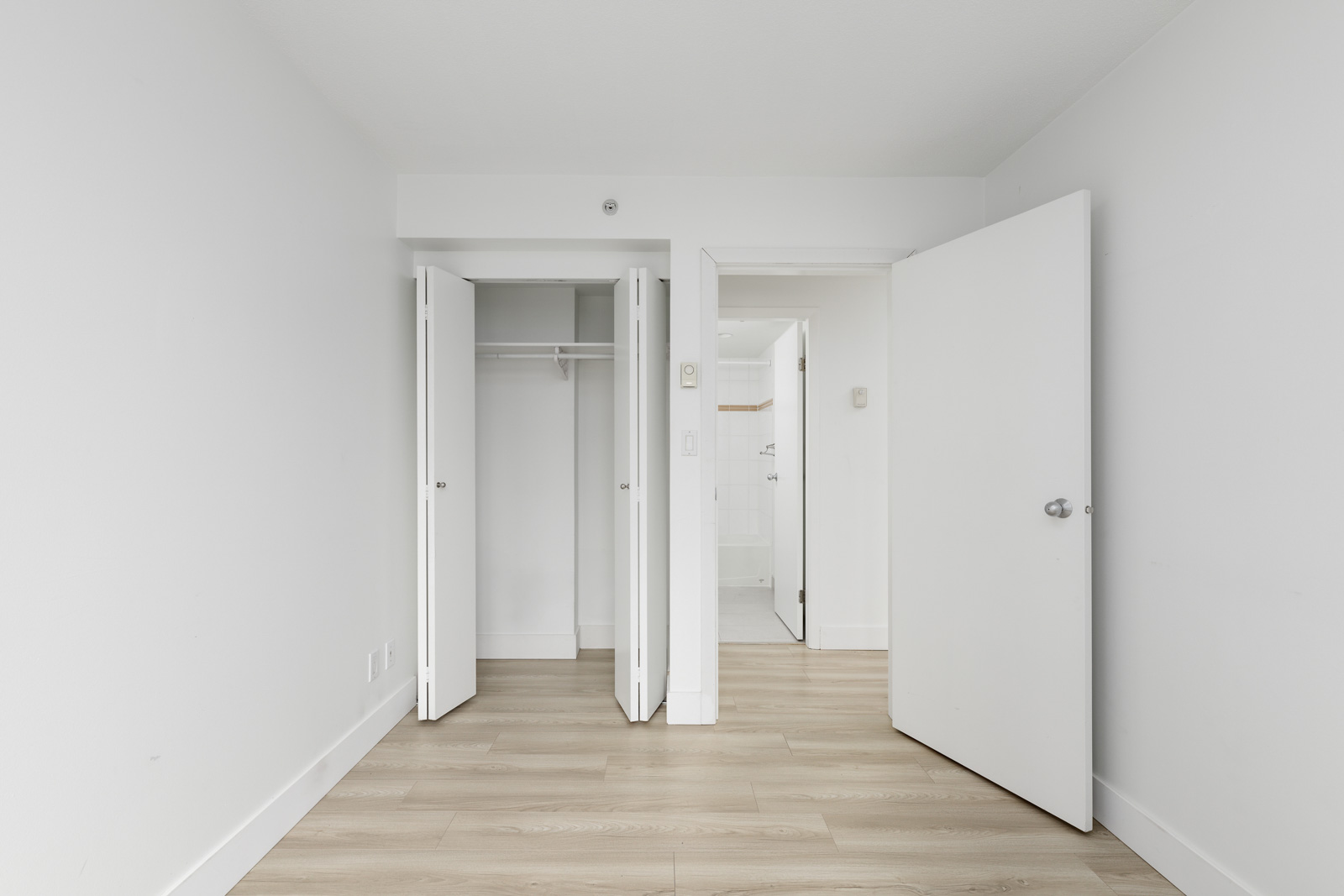 Empty room with light wood flooring, white walls, an open closet with double doors, and an open door leading to another room with white tile flooring.