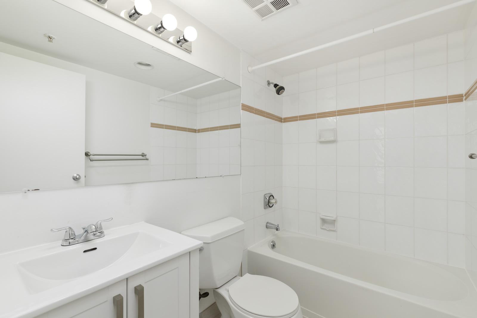 White bathroom with a sink, toilet, bathtub, large mirror, white tiled walls, and overhead lighting.