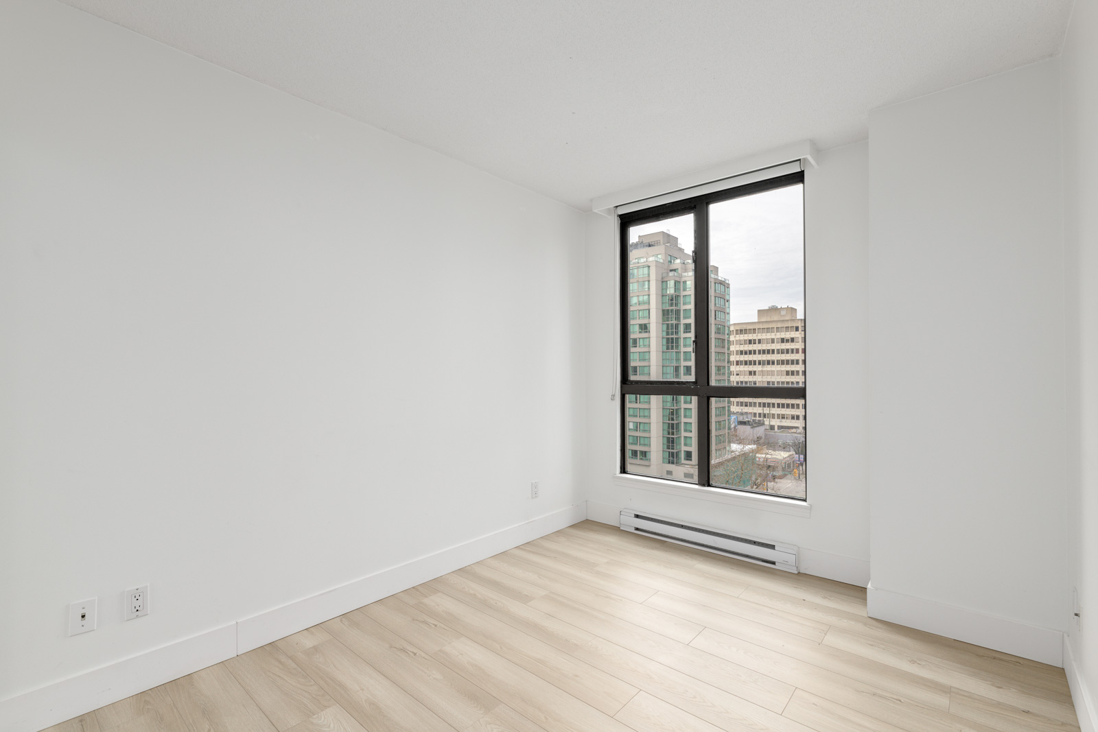 Empty room with light wood flooring, white walls, and a large window showing a cityscape with tall buildings outside.
