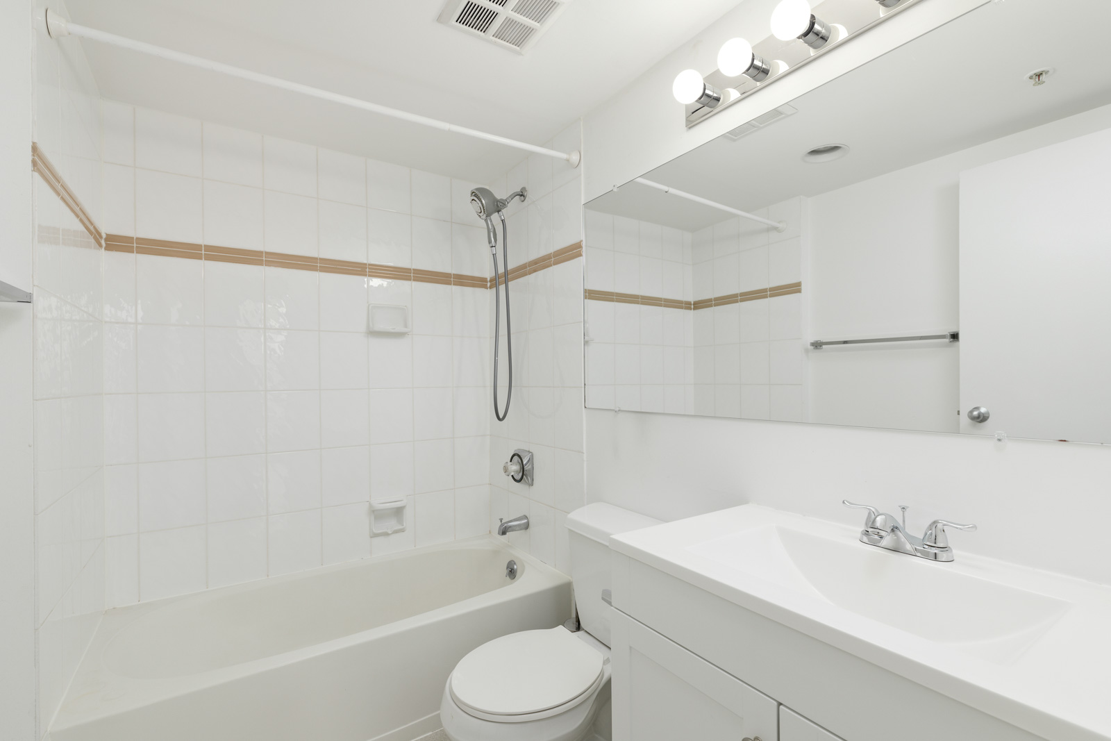 White bathroom with bathtub-shower combo, toilet, large mirror, vanity with sink, and overhead lights; walls and tiles are mostly white with a beige accent stripe.