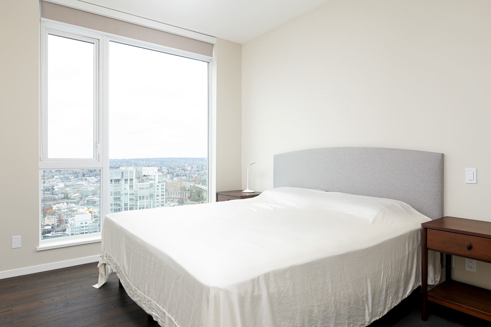 Minimalist bedroom with a neatly made bed, two wooden nightstands, a lamp, and a large window overlooking a cityscape.