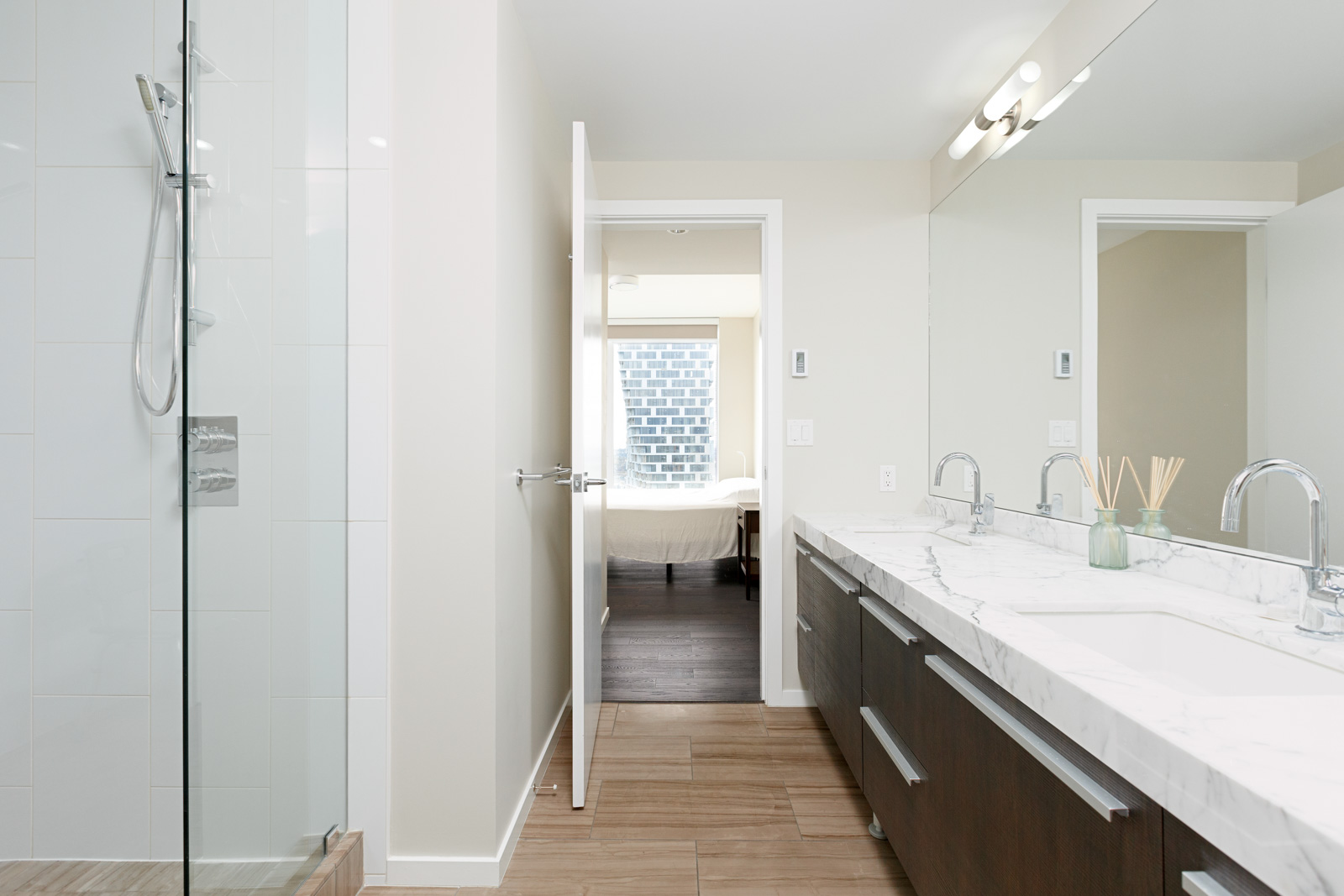 Modern bathroom with a glass shower, double sinks with marble countertop, large mirror, and view into a bedroom through an open door.