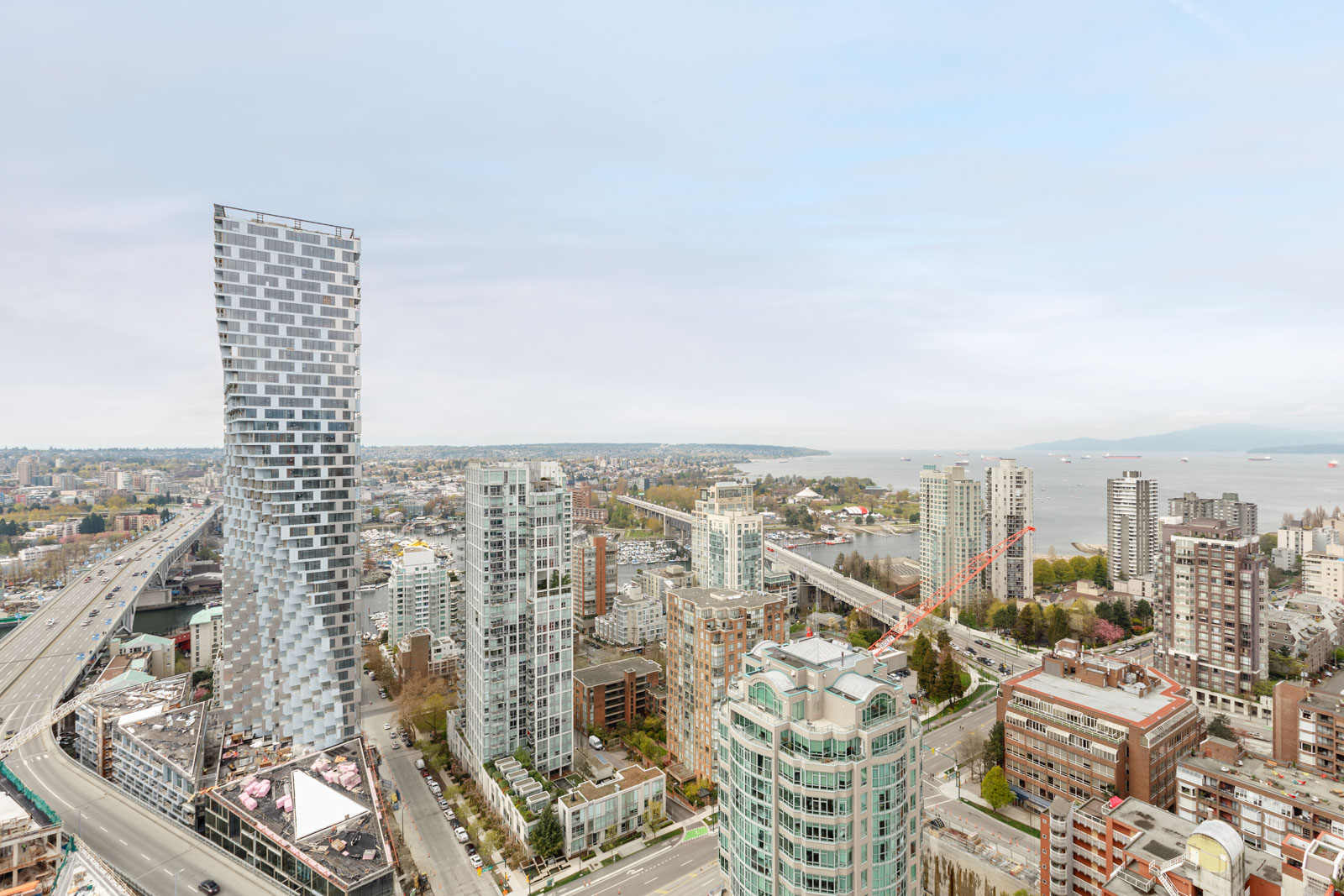 Aerial view of a modern cityscape with high-rise buildings, a unique twisted tower, roads, and a body of water in the background under a cloudy sky.