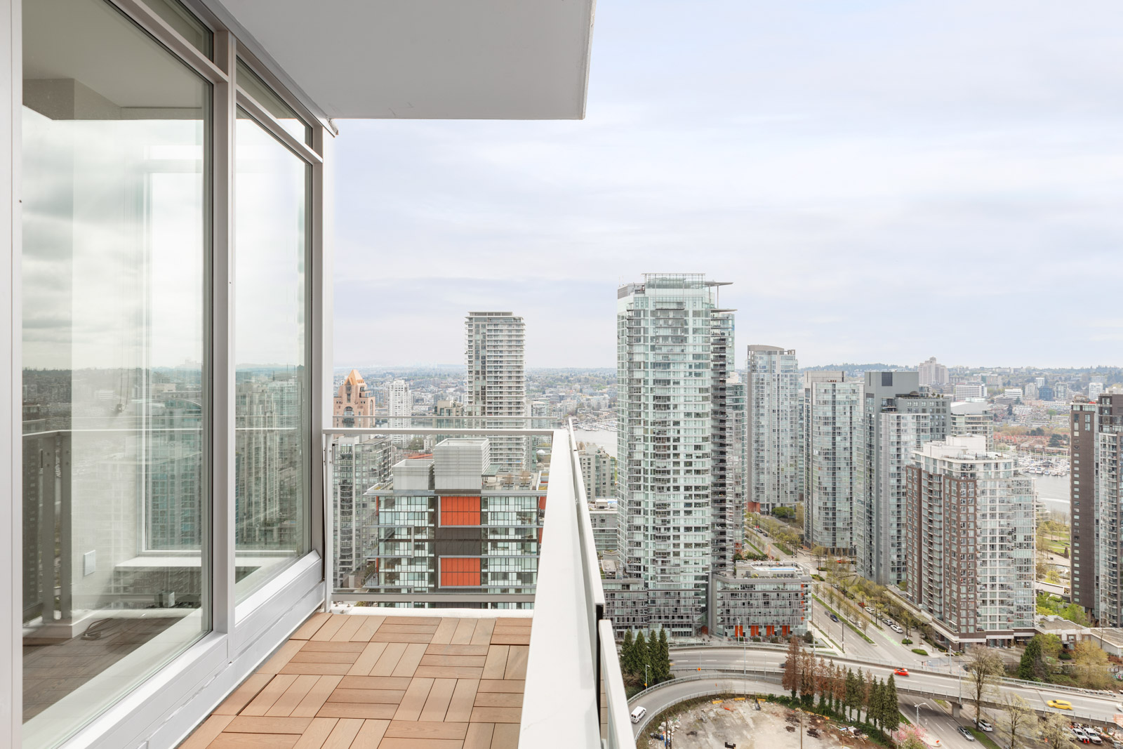 High-rise apartment balcony with glass railing overlooking a cityscape with tall buildings, roads, and a cloudy sky.