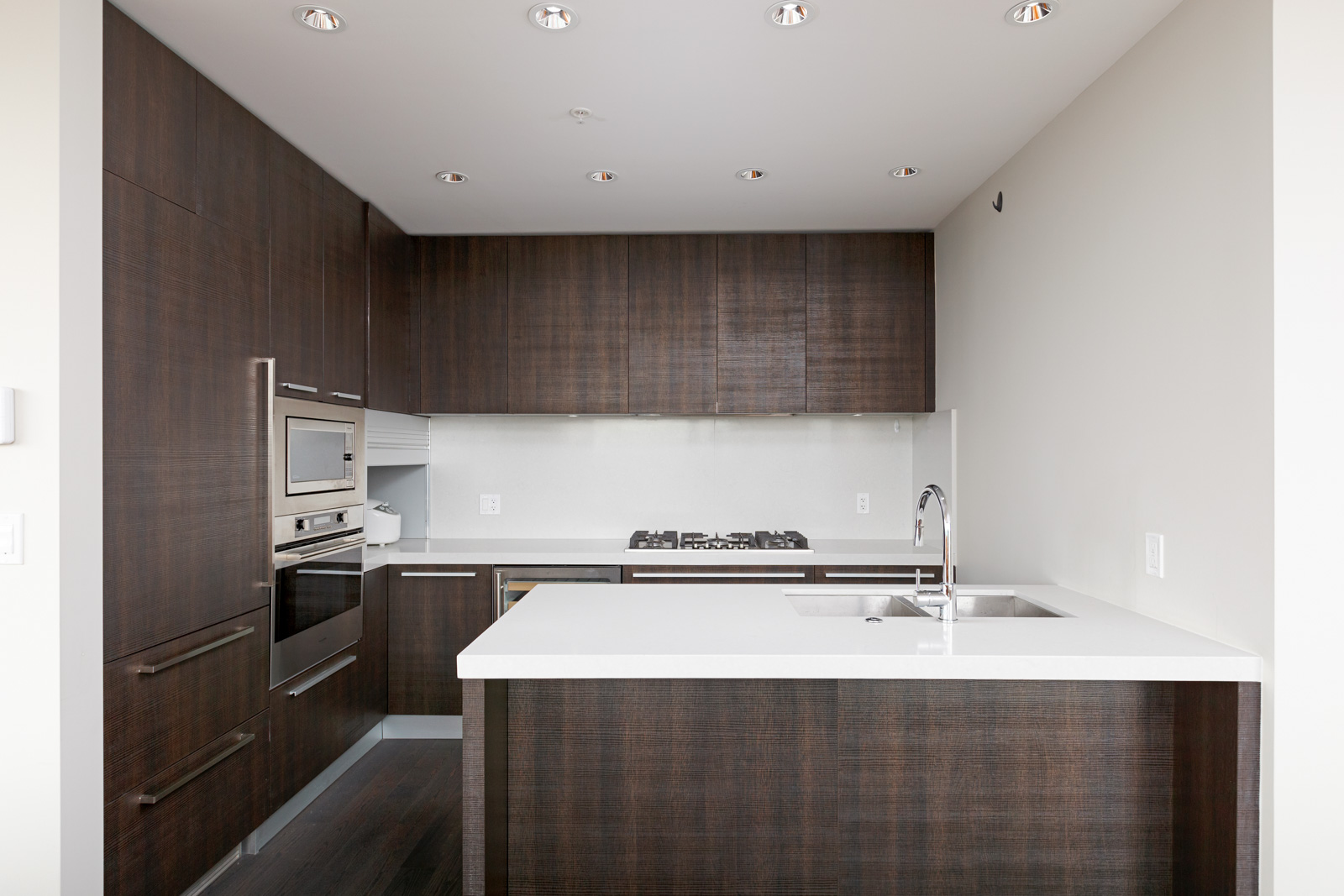 Modern kitchen with dark wood cabinets, built-in stainless steel appliances, white countertops, gas cooktop, and a sink on the island under recessed ceiling lights.