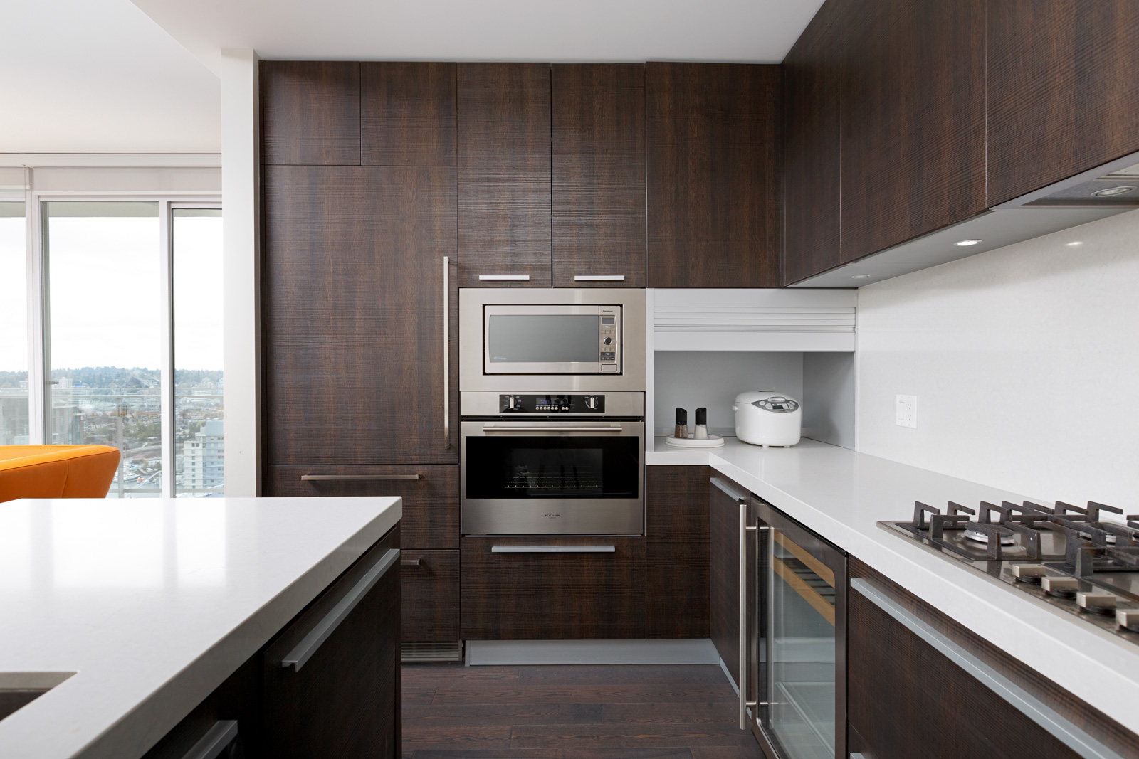 Modern kitchen with dark wood cabinets, built-in stainless steel appliances, a white countertop, gas stove, and a rice cooker on the counter. Large windows provide natural light.