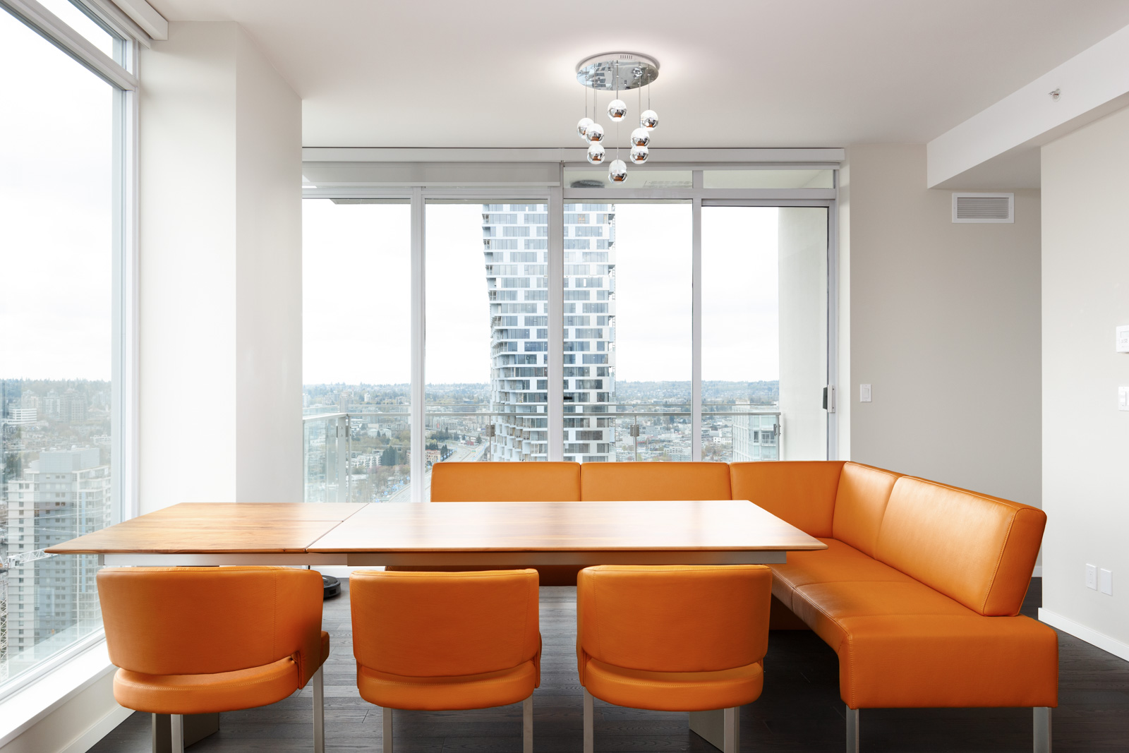 Modern dining area with orange chairs and an L-shaped bench, large windows, and a view of a cityscape with a unique high-rise building.