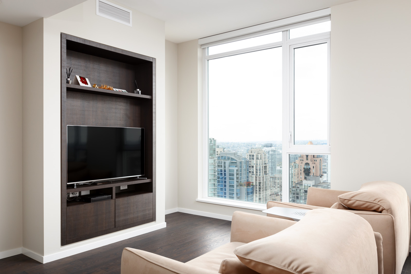 Modern living room with beige sofas, a wall-mounted TV in a dark wood entertainment unit, large window, and cityscape view.