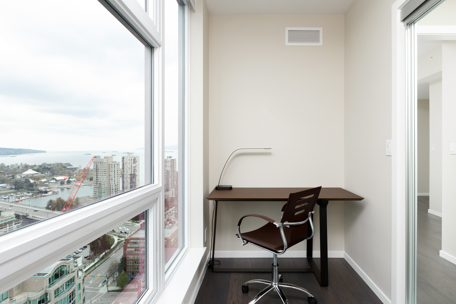 A small, modern home office with a desk, chair, and lamp is positioned by large windows overlooking a cityscape and waterfront.