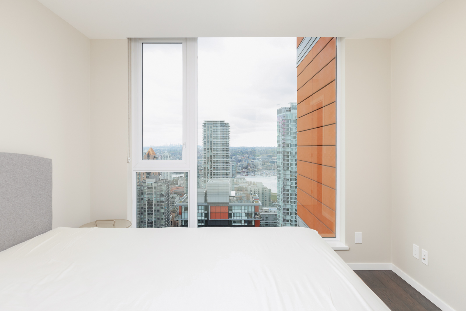 A modern bedroom with a large window overlooking city buildings and a waterfront; the room has white walls, a bed, and dark wood flooring.