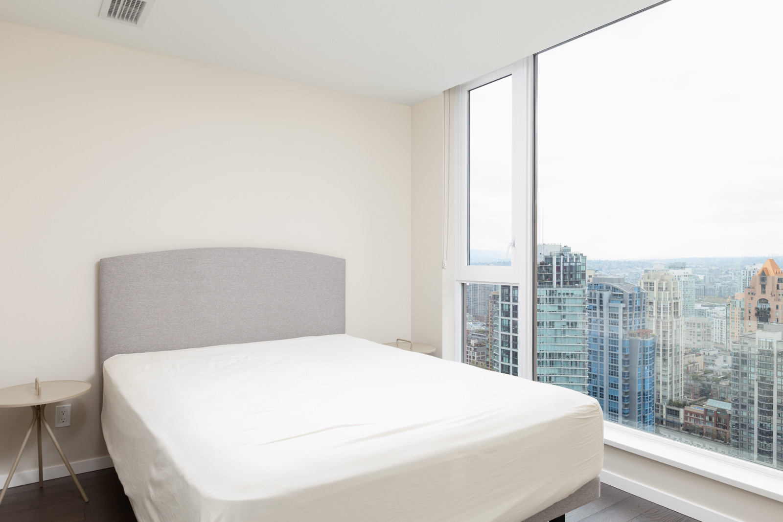 Minimalist bedroom with a bed, small round side table, and floor-to-ceiling window overlooking city skyscrapers.