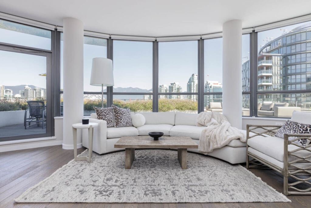 Modern living room with large curved windows, a white sectional sofa, patterned rug, and wooden coffee table. City buildings and mountains visible through the windows.