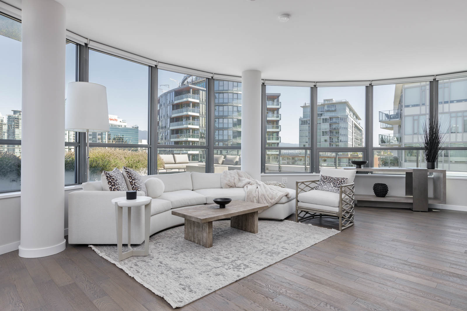 Modern living room with a white sectional sofa, armchair, wooden coffee table, rug, and large windows overlooking urban buildings.