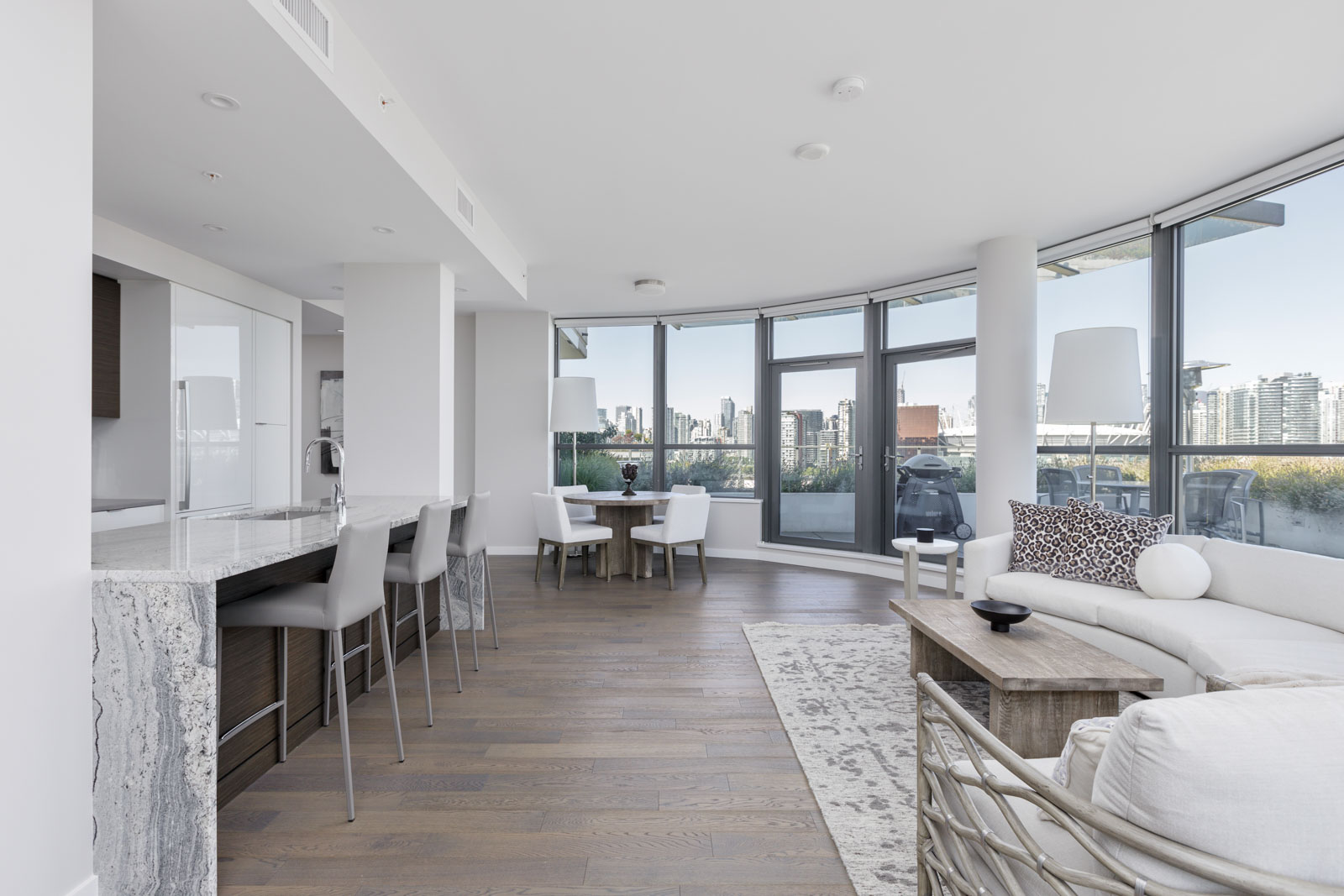 Modern open-plan living area with kitchen, dining table, and curved windows overlooking a cityscape; light furnishings and wooden flooring throughout the space.