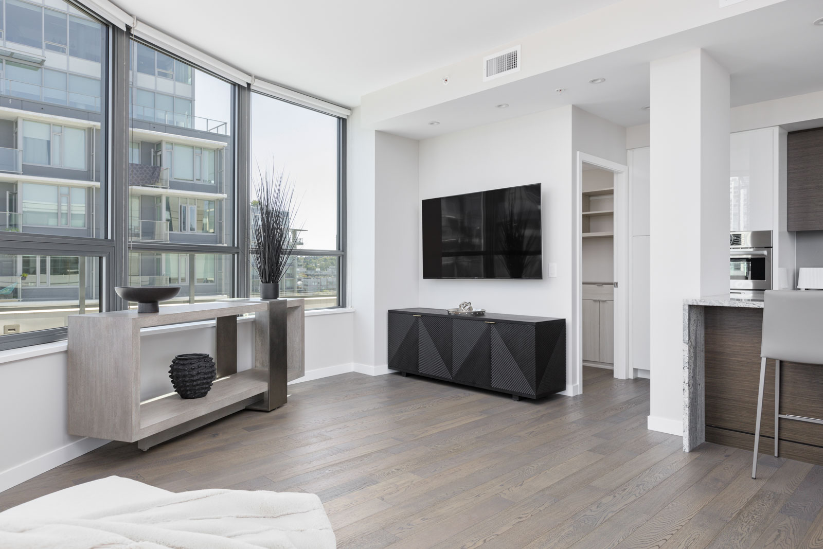 Modern living room with large windows, wall-mounted TV, minimalistic furniture, light wood flooring, and an open kitchen area in a high-rise apartment.