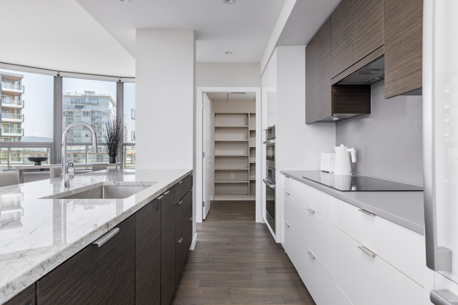 Modern kitchen with dark and white cabinets, marble island, stainless steel sink, and view of a walk-in pantry; large windows show neighboring buildings outside.
