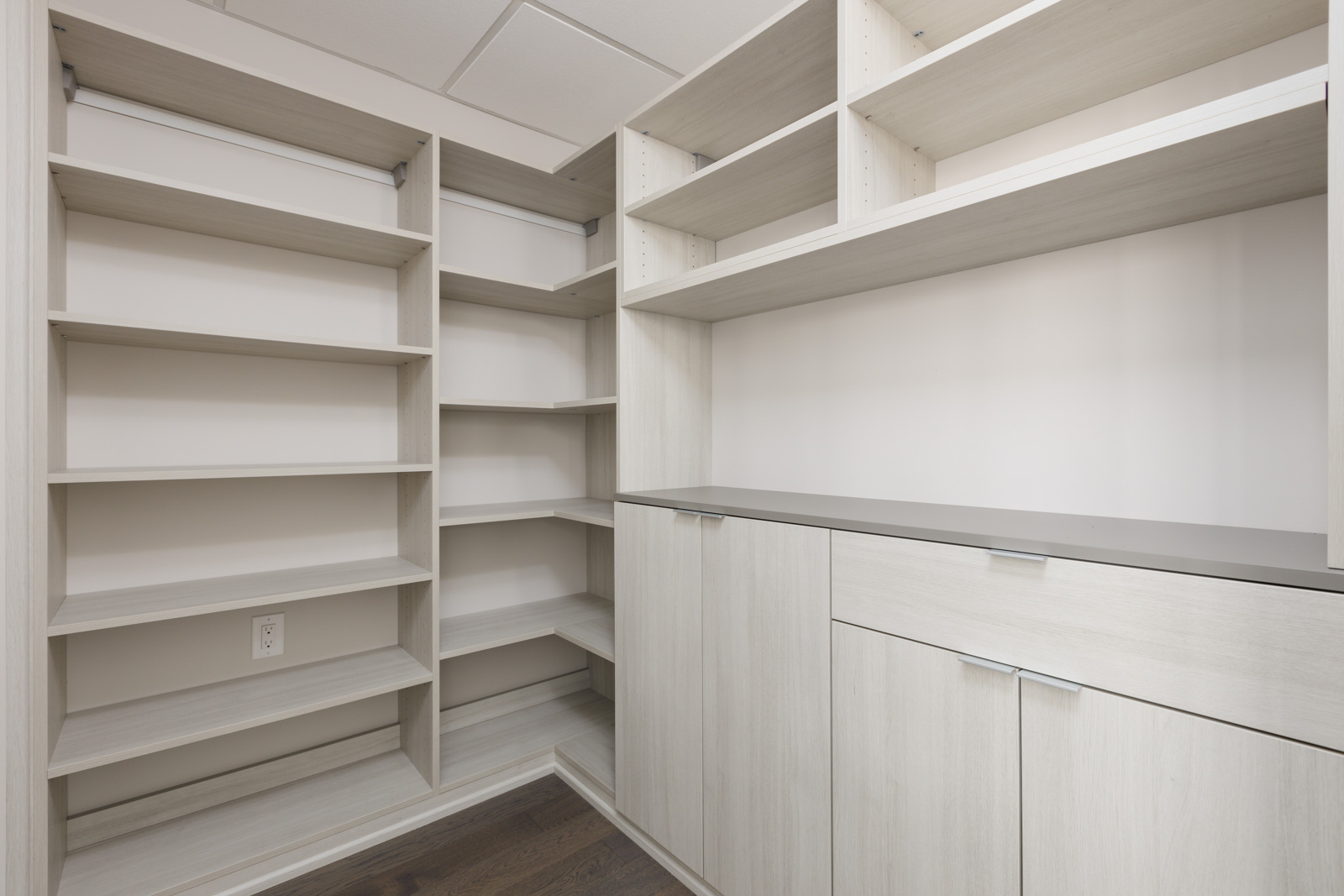 Empty walk-in closet with light wood shelves, cabinets, and a gray countertop, featuring a corner layout and white walls.