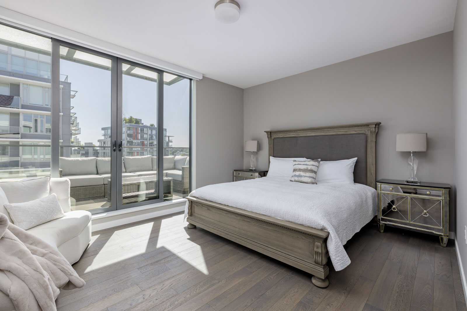 Modern bedroom with large glass doors, a gray bed with white bedding, mirrored nightstands, and an armchair; city buildings visible outside on the balcony.