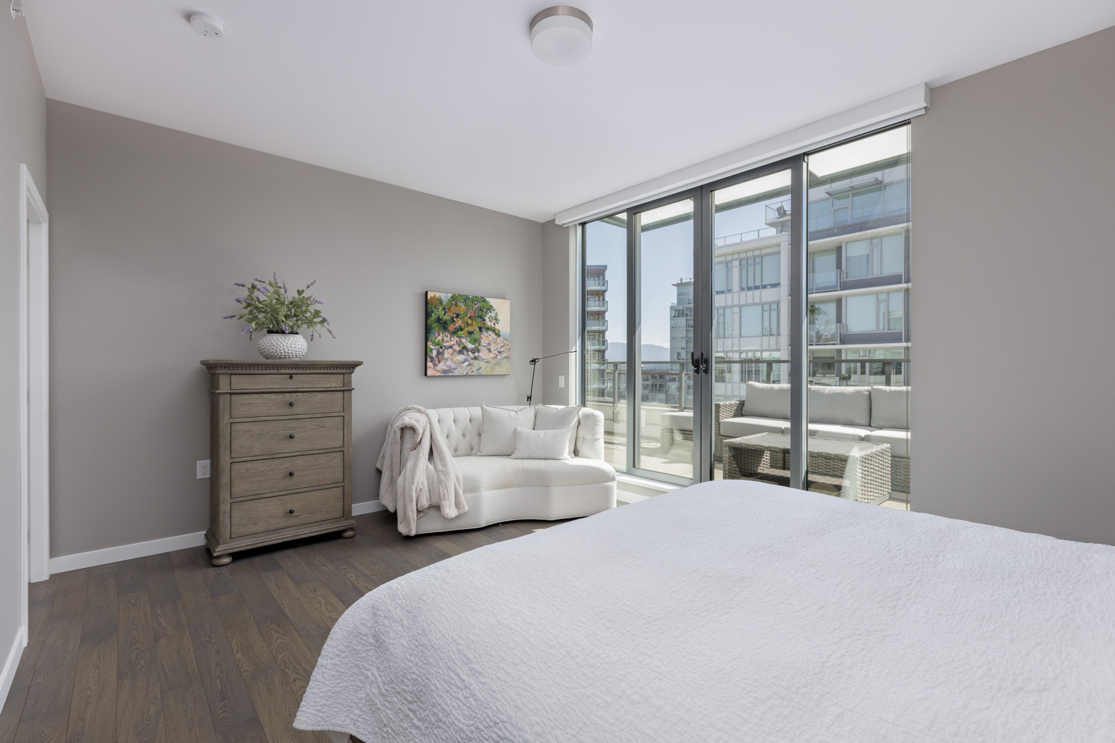 A modern bedroom with a large bed, wooden dresser, white chaise lounge, wall art, and sliding glass doors leading to a balcony with outdoor seating.